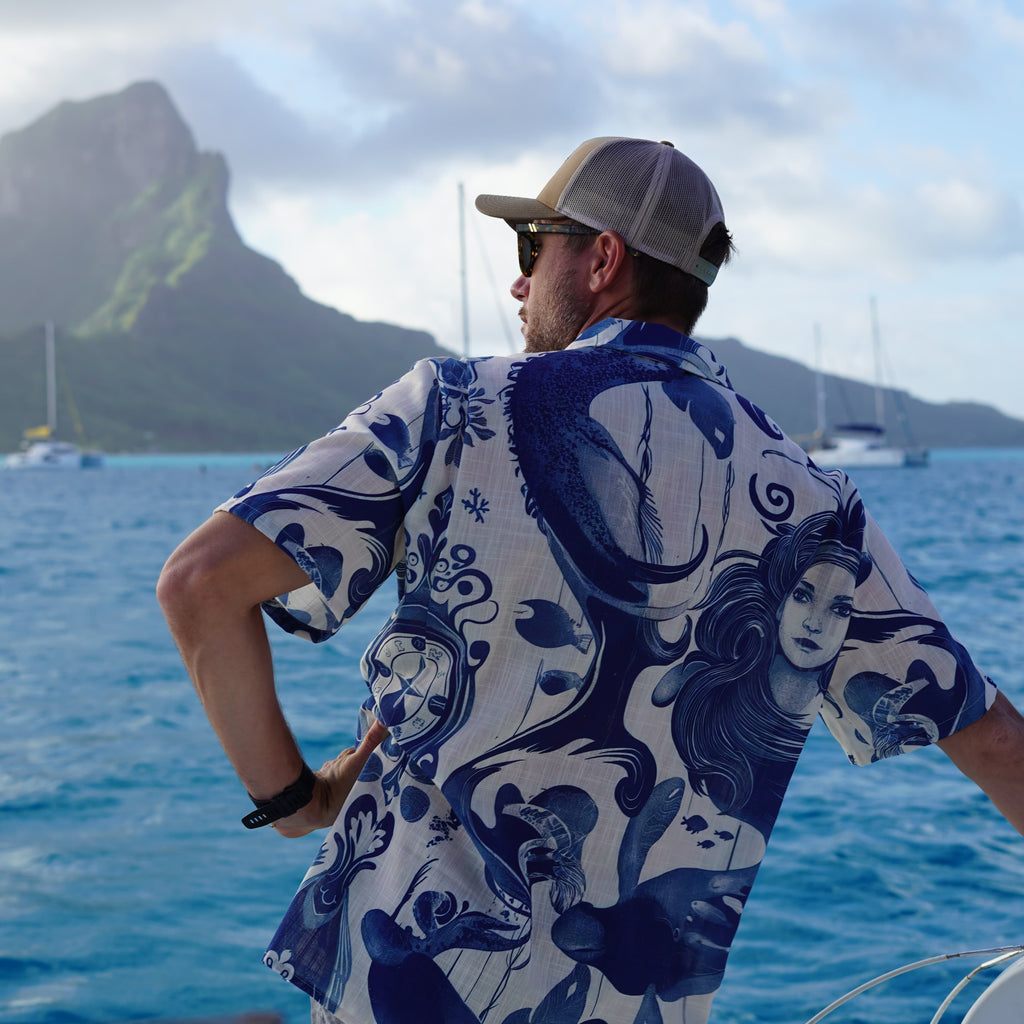 A man on a boat wearing a blue and white patterned short-sleeve button-up shirt, sunglasses, and a baseball cap, looking out over blue water toward green mountains under a cloudy sky.