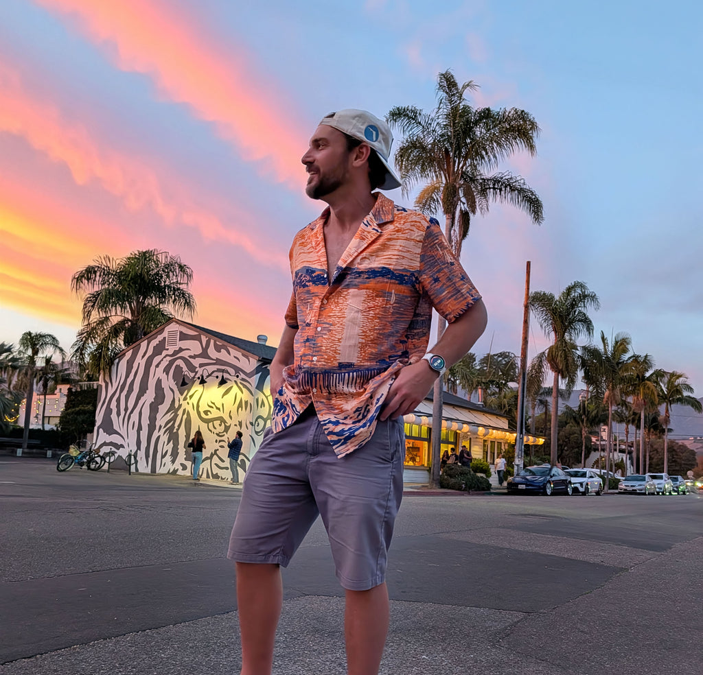A man wearing the Last Light at Stearns shirt and shorts on a palm-lined street under a pink sunset.